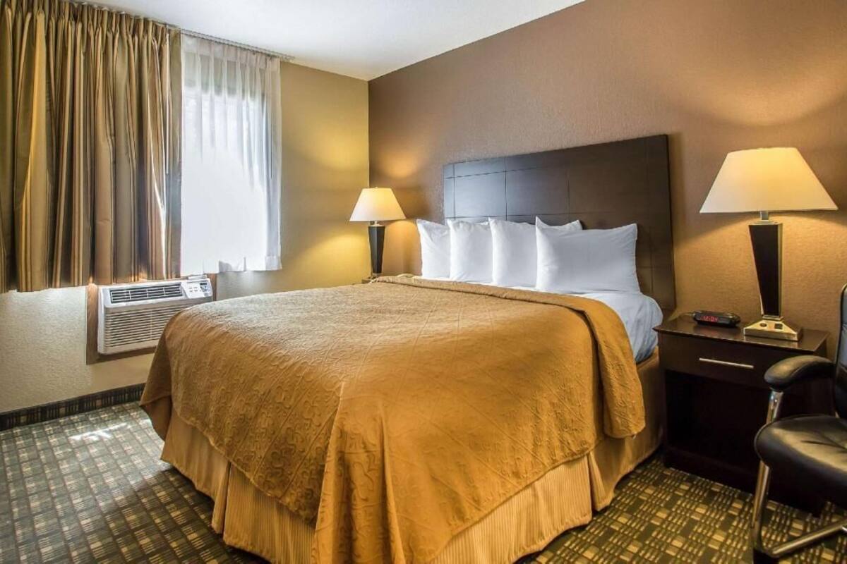 A hotel room with a large bed, two bedside lamps, a chair, and an air conditioning unit.