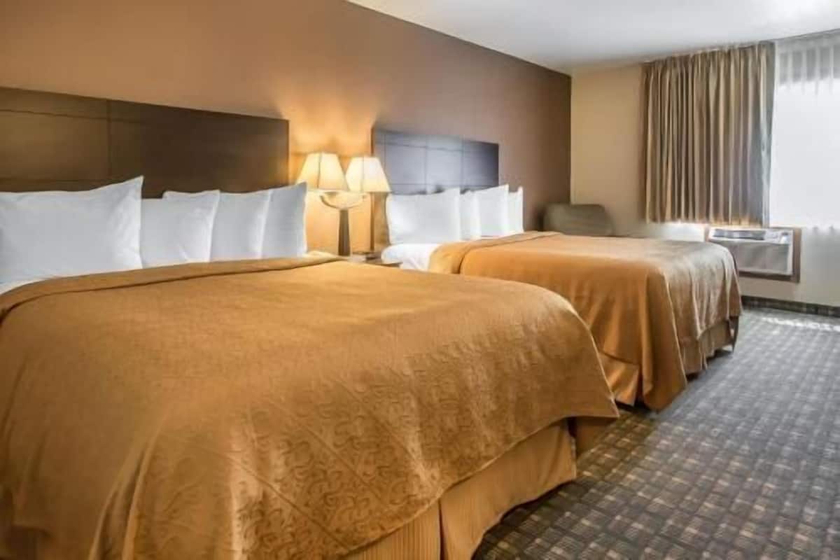 A hotel room with two beds, a headboard, a lamp, a chair, and a window with curtains.