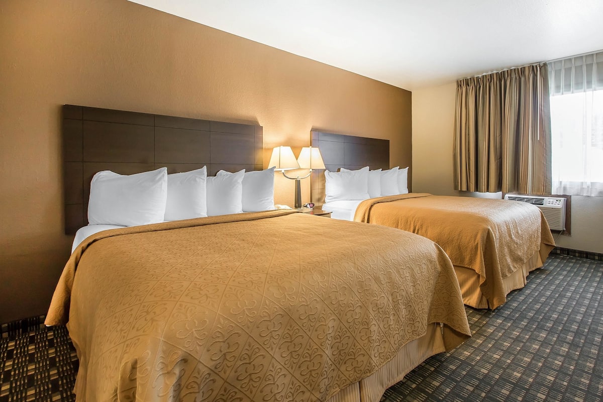 A hotel room with two beds, a headboard, a lamp, an air conditioning unit, and a window with curtains.