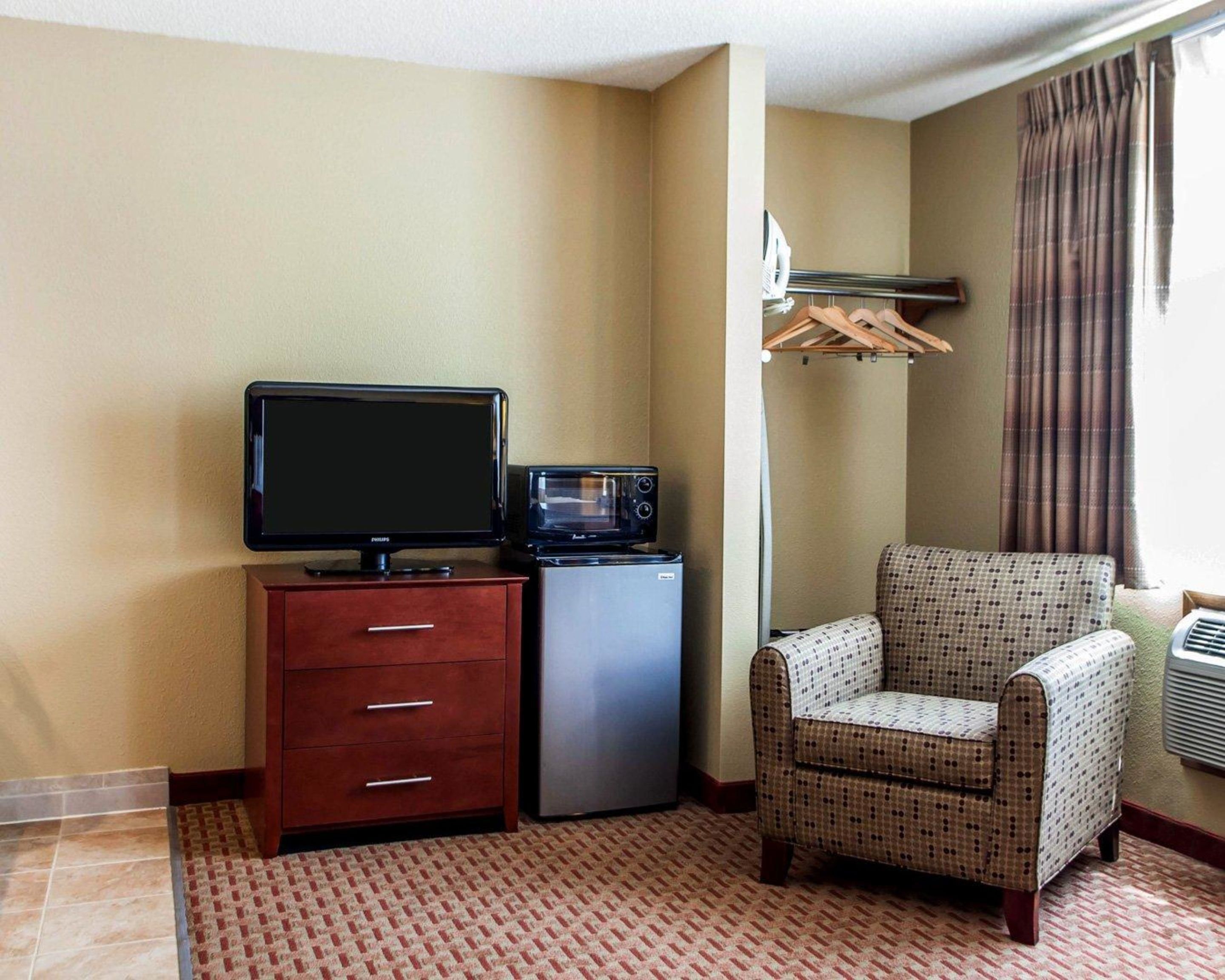 A hotel room with a television, a microwave, a small refrigerator, a patterned armchair, and a clothing rack.