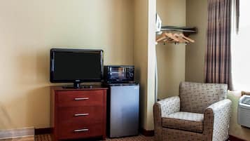 A hotel room with a television, a microwave, a small refrigerator, a patterned armchair, and a clothing rack.