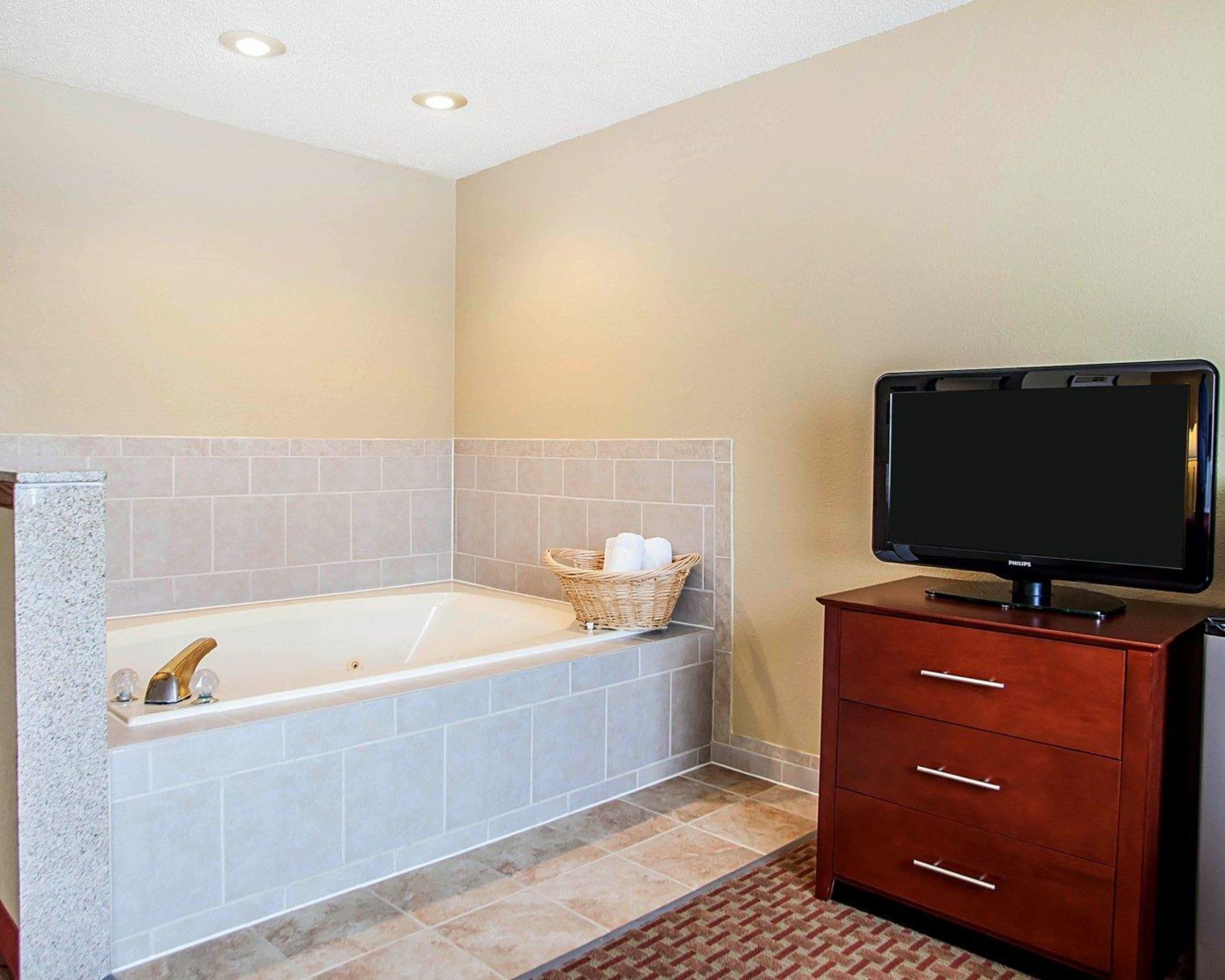 Un baño con bañera, un televisor sobre un mueble de madera y una cesta de mimbre con toallas.