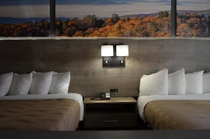 A hotel room with two beds, a nightstand with a phone, and a view of autumn foliage through large windows. - Quality Inn & Suites Ankeny - Des Moines (Ankeny)