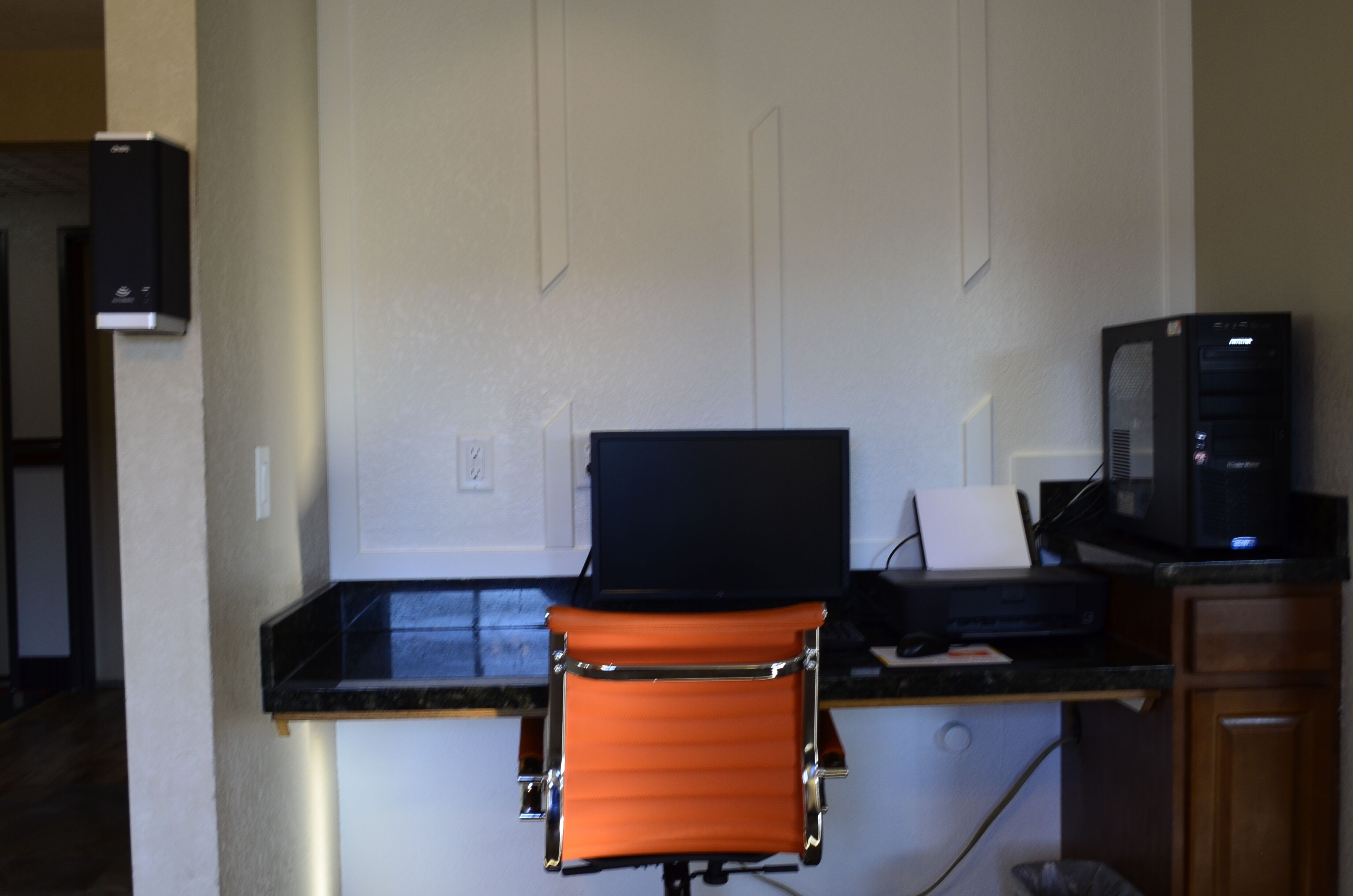 Un petit espace de bureau comprenant un bureau noir, un écran d’ordinateur et une chaise orange.
