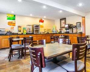 Un comedor de hotel con una mesa grande y varias sillas, una zona de cocina con electrodomésticos y un televisor montado en la pared.