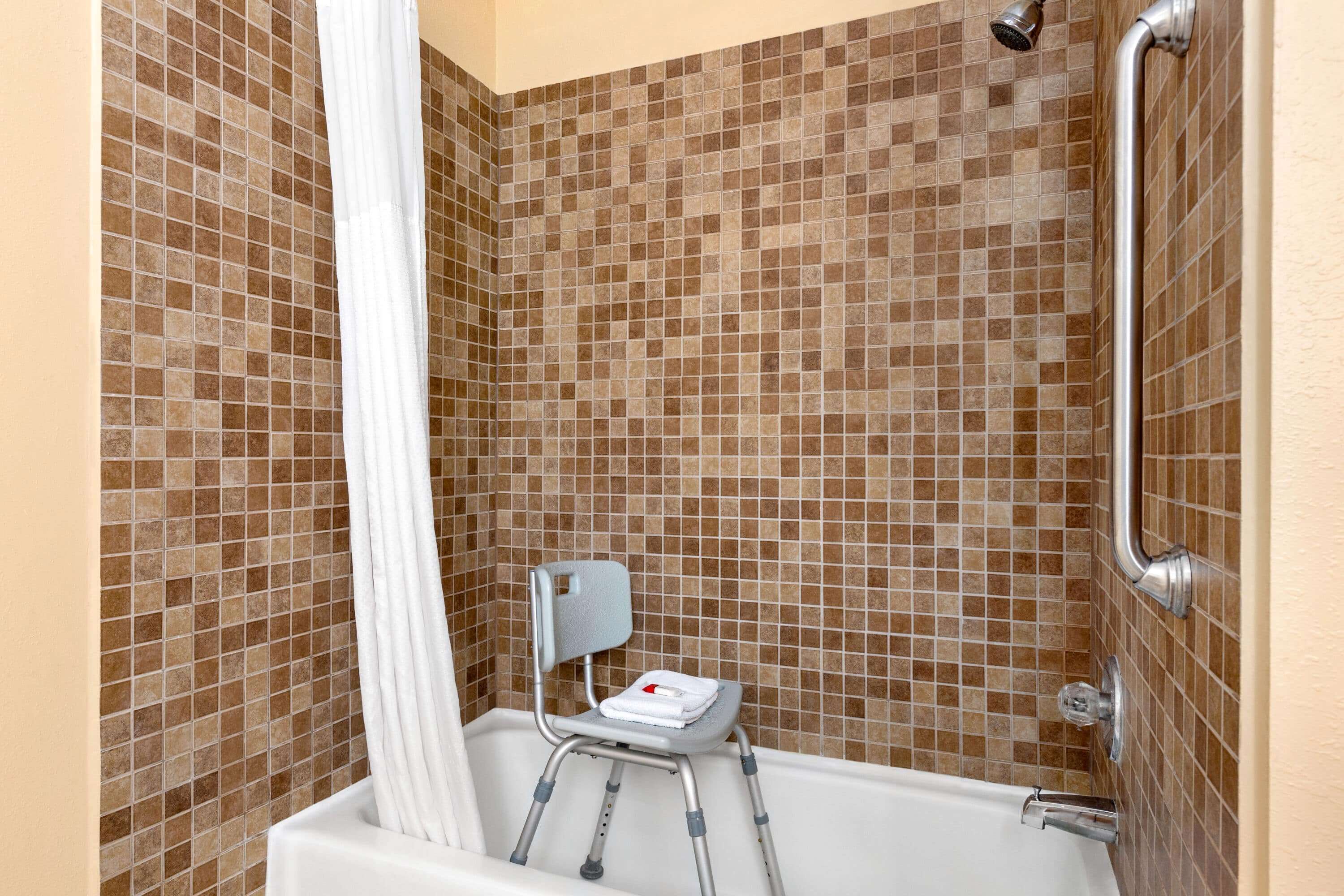 A bathroom with a shower, tiled walls, a white bathtub, and a small chair with a towel.