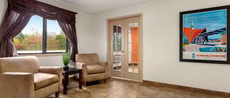 A room with two beige armchairs, a small table with a plant, a window with curtains, and a door leading outside.