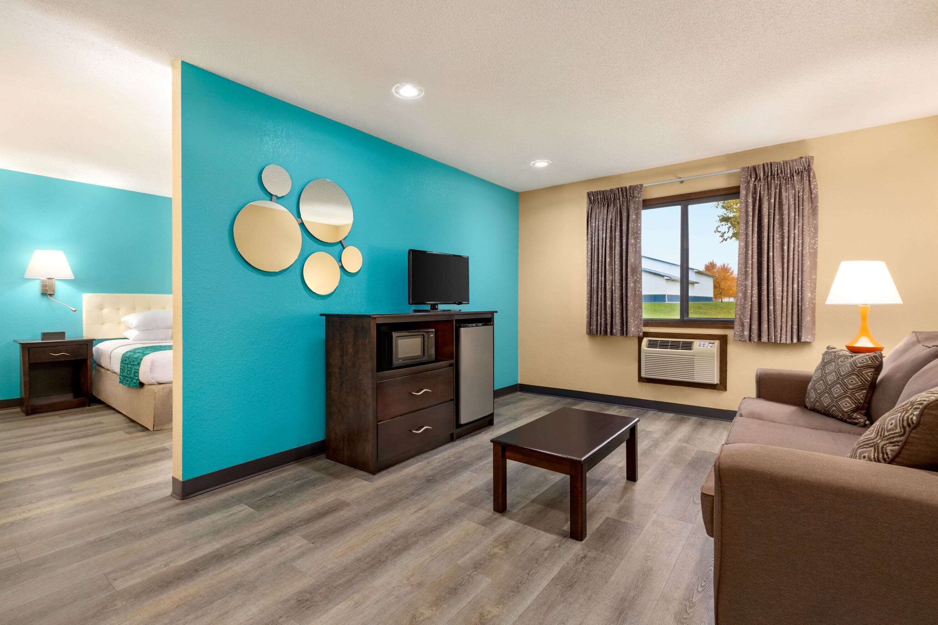 Une chambre d’hôtel avec un mur d’accent de couleur turquoise, un lit, une télévision, une petite table, un canapé et une fenêtre avec des rideaux.