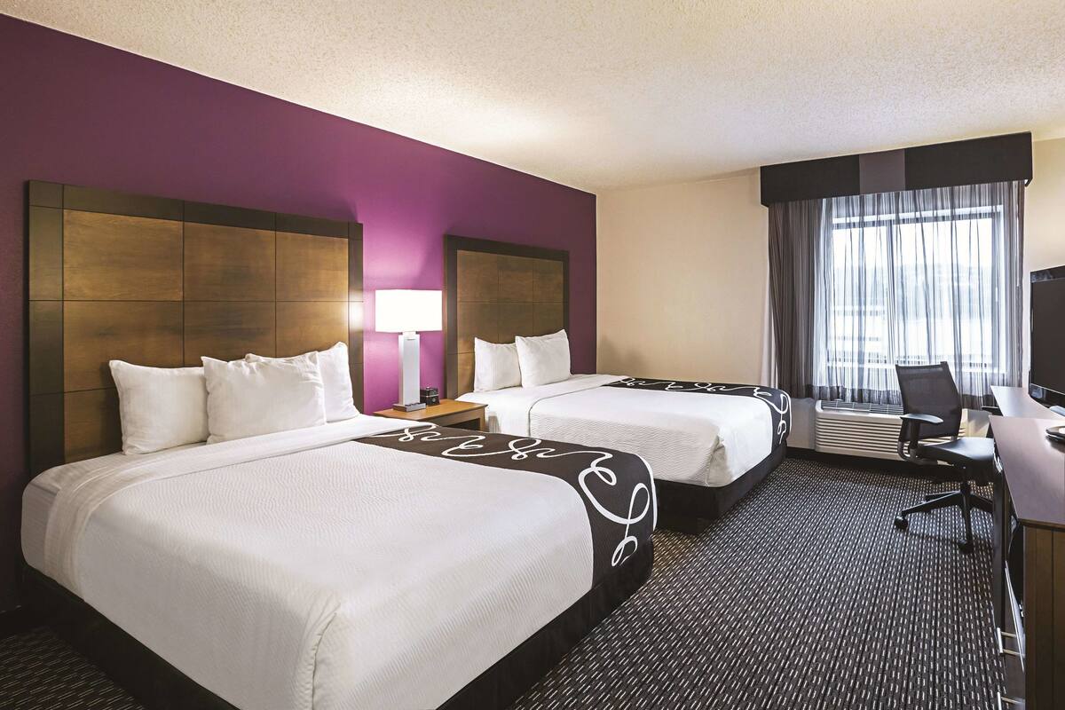 A hotel room with two beds, a desk, a chair, a lamp, and a large window with curtains.