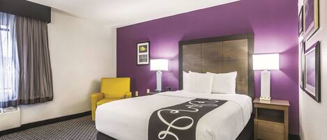Habitación de hotel con una cama grande, un sillón amarillo, dos mesitas de noche con lámparas y una ventana con cortinas.