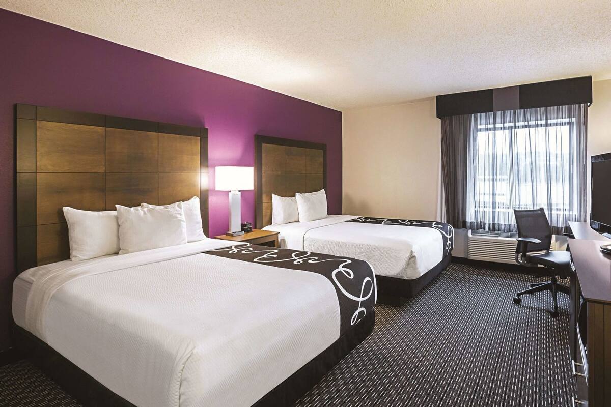 A hotel room with two beds, a desk, a chair, and a large window with curtains.