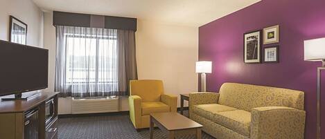Een hotelkamer met een televisie, twee stoelen, een bank, een lamp en een raam met gordijnen.