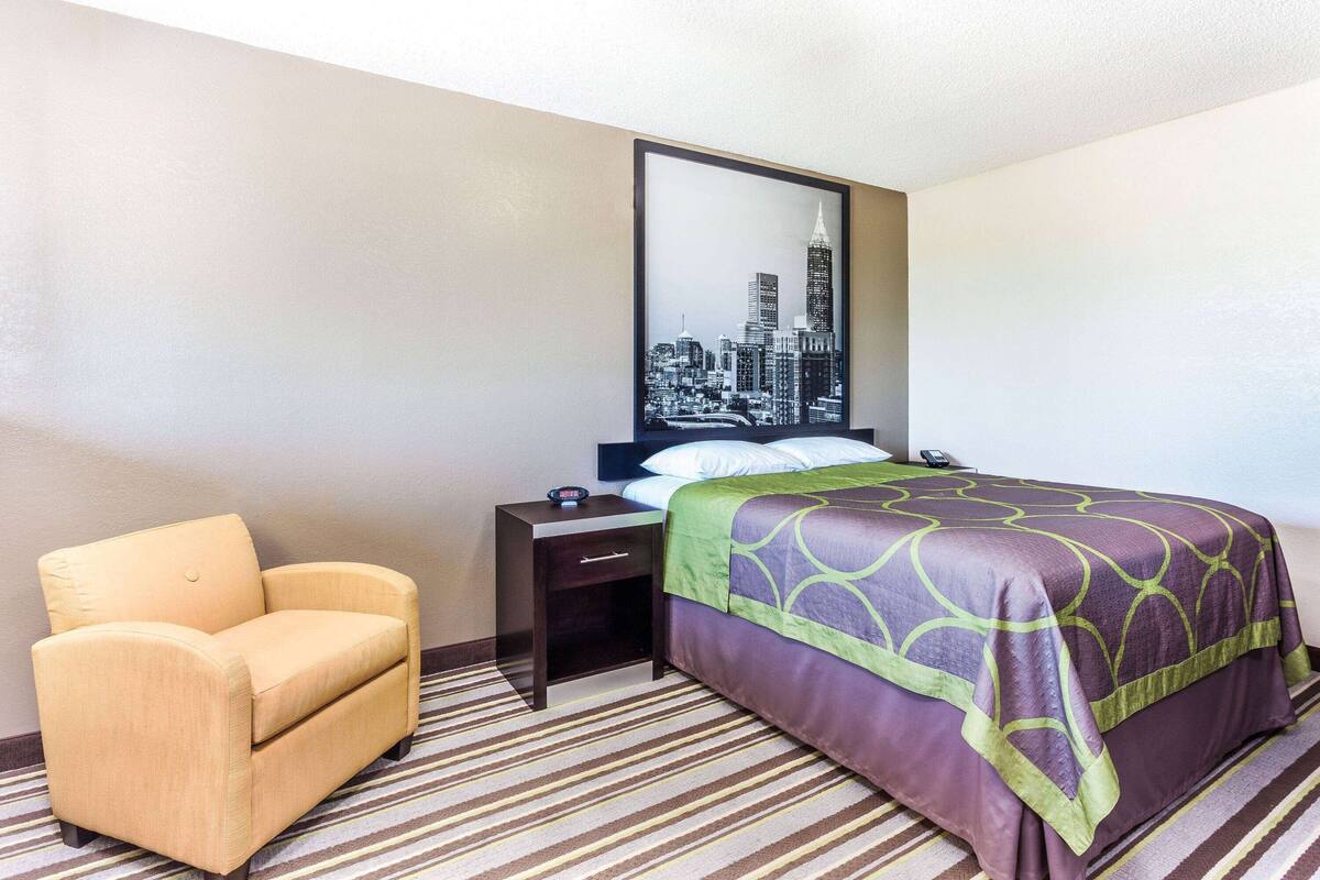 A hotel room with a bed, a chair, a nightstand, and a cityscape picture on the wall.