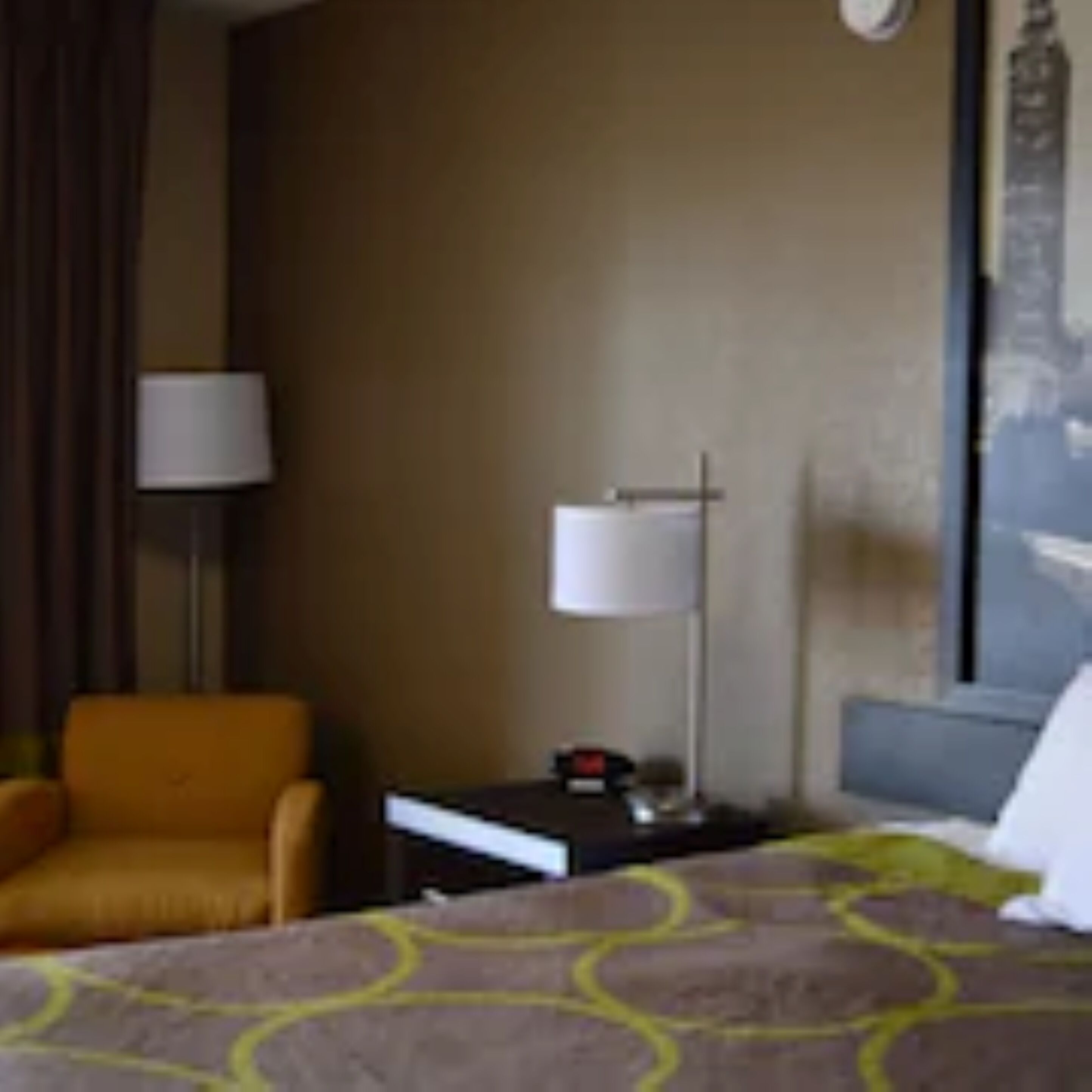 A hotel room with a bed, a nightstand, a lamp, a chair, and a wall with a painting.