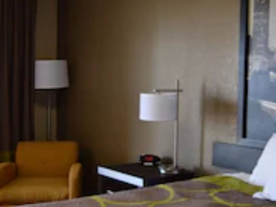 A hotel room with a bed, a nightstand, a lamp, a chair, and a wall with a painting.