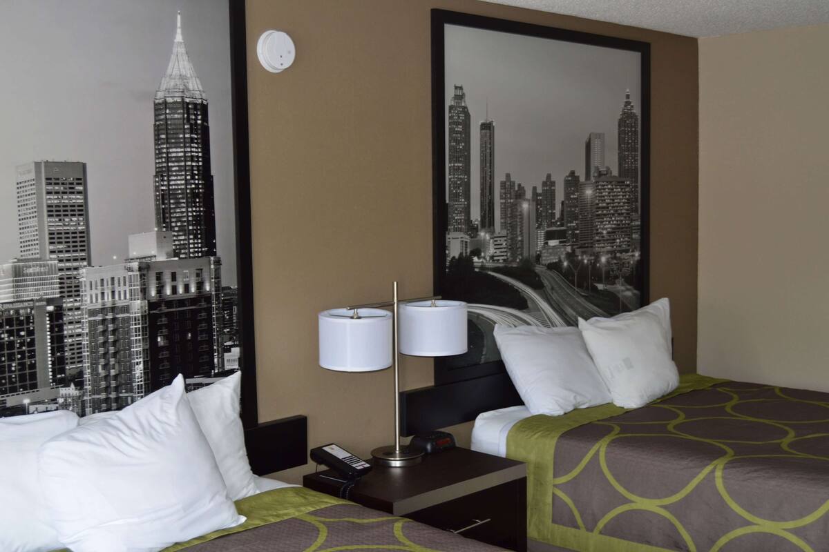 A hotel room with a bed, pillows, a nightstand with a lamp, a phone, and a cityscape painting on the wall.