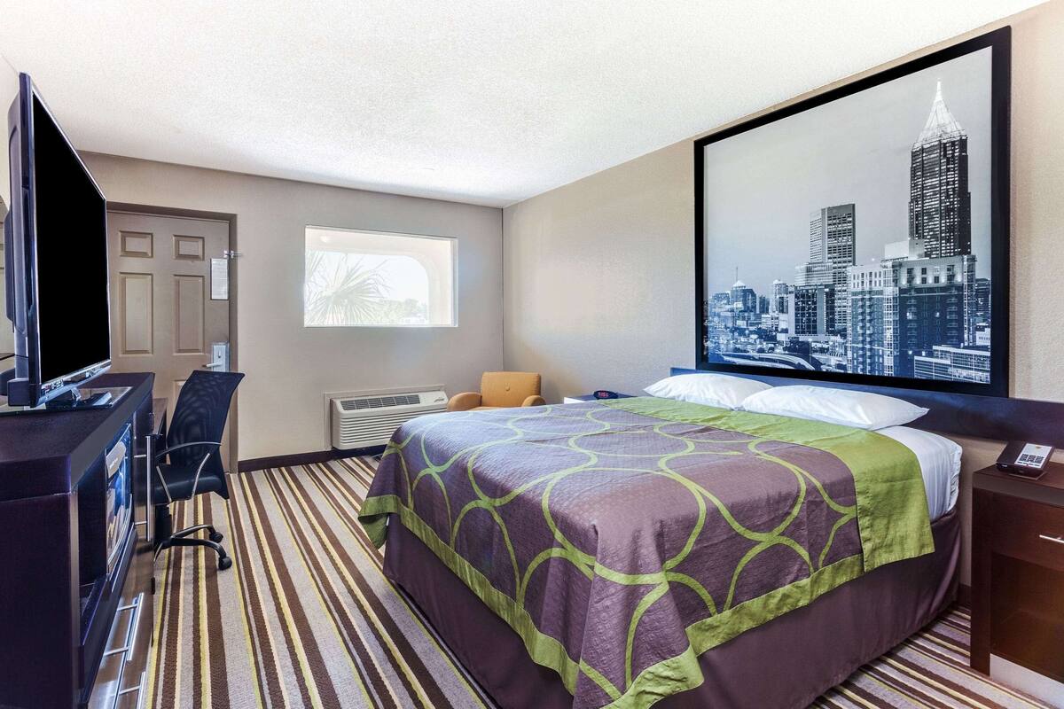 A hotel room with a bed, a desk with a computer, a chair, a window with a cityscape view, and a large framed picture on the wall.