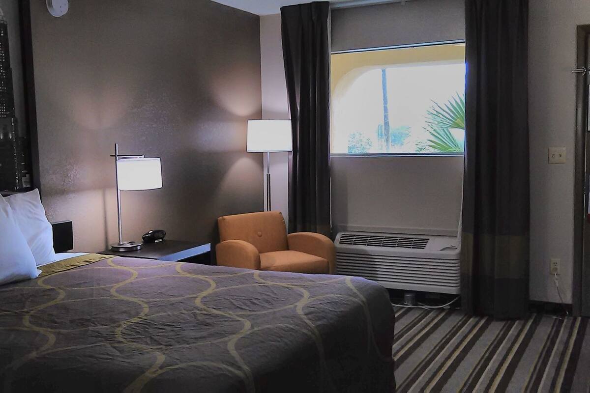 A hotel room with a bed, a chair, a desk, a window with curtains, and an air conditioning unit.