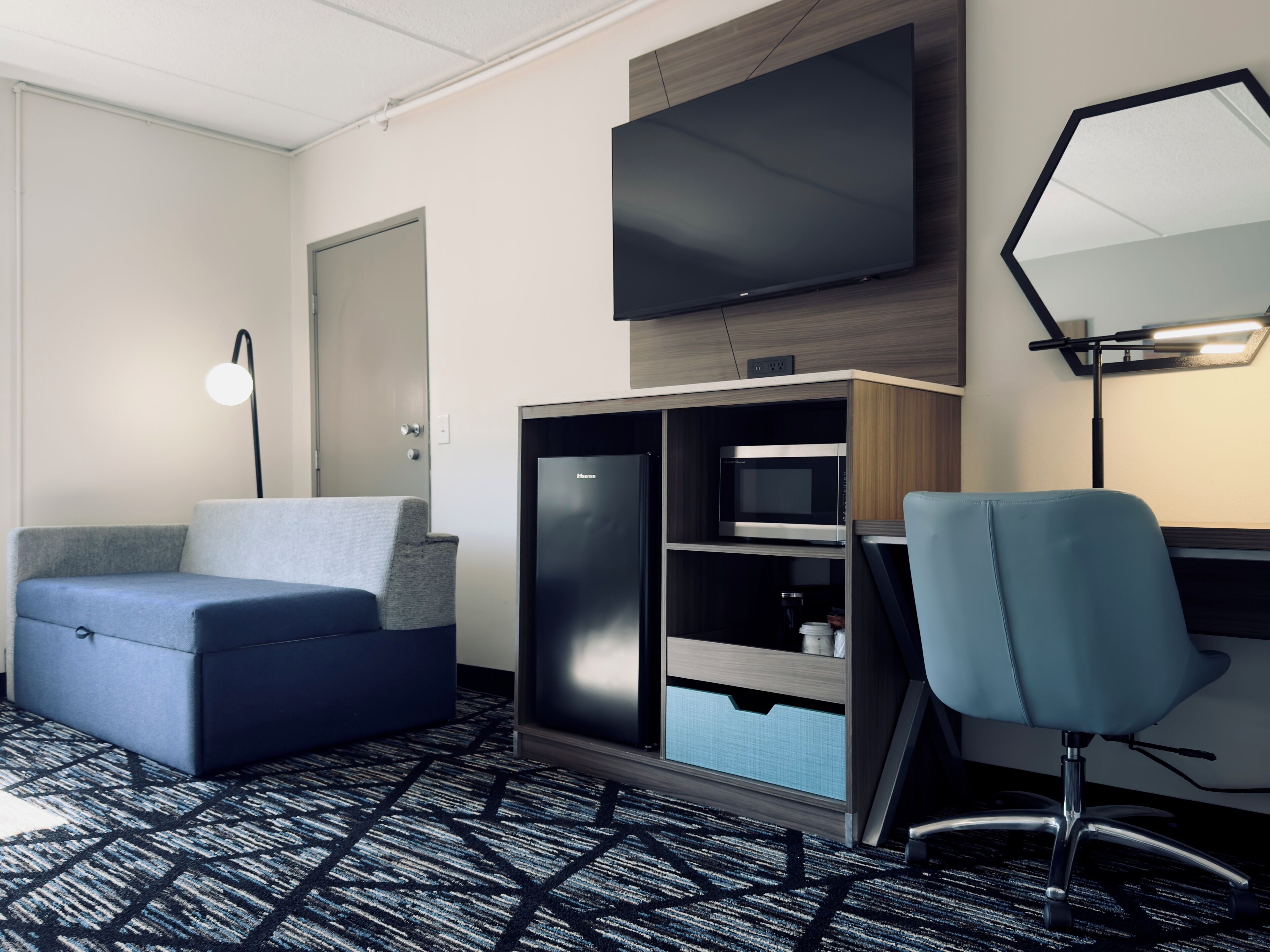 Habitación de hotel con sofá, escritorio y silla, televisor de pantalla plana montado en la pared, microondas y un mueble con cajones.