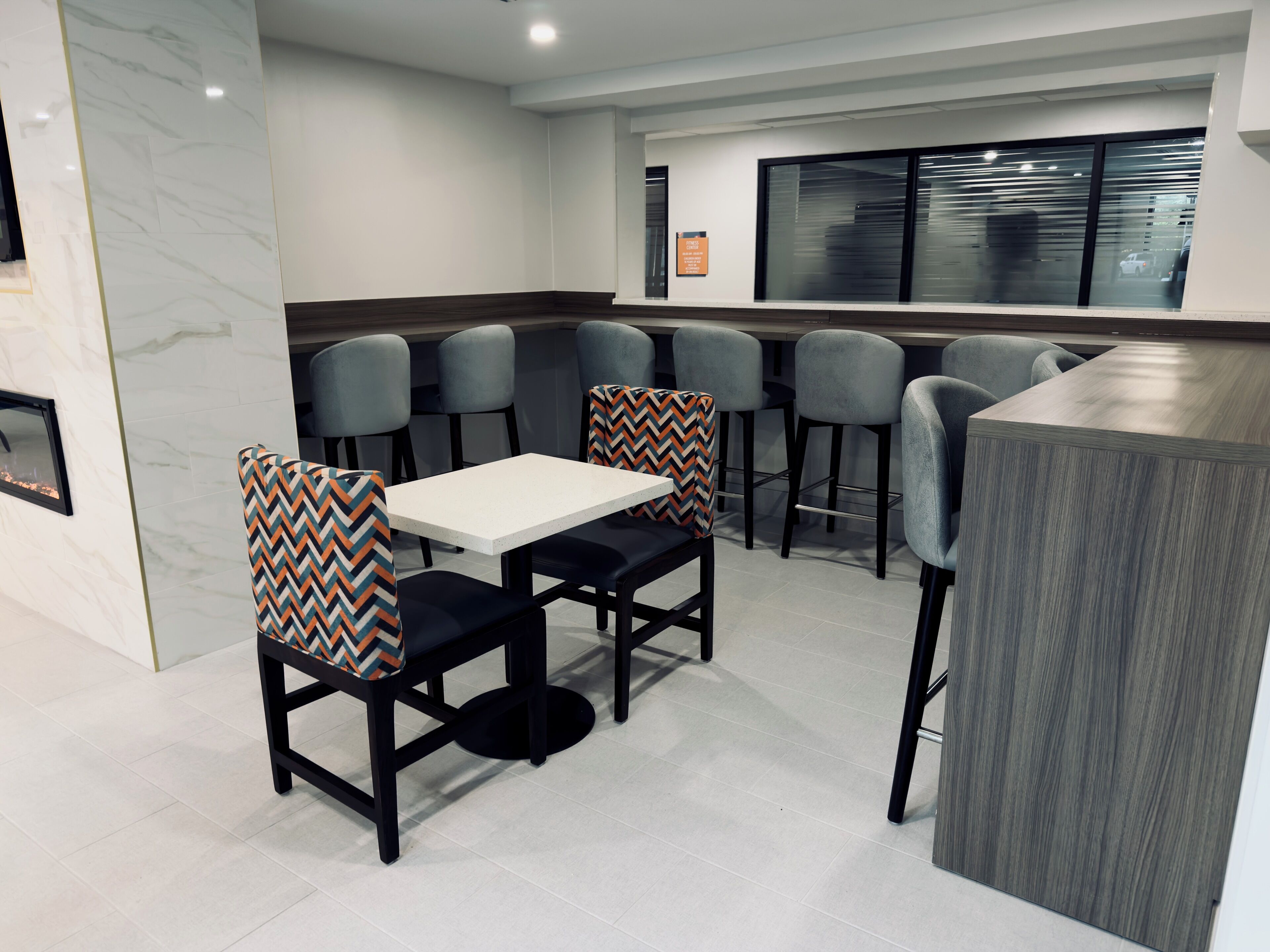 Un comedor moderno con una mesa blanca y sillas negras, una zona de bar con mostrador gris, y un ventanal con persianas.