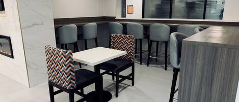Un comedor moderno con una mesa blanca y sillas negras, una zona de bar con mostrador gris, y un ventanal con persianas.