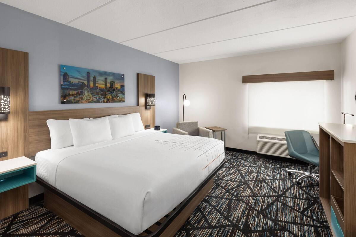 A hotel room with a large bed, a desk, a chair, and a cityscape painting on the wall.