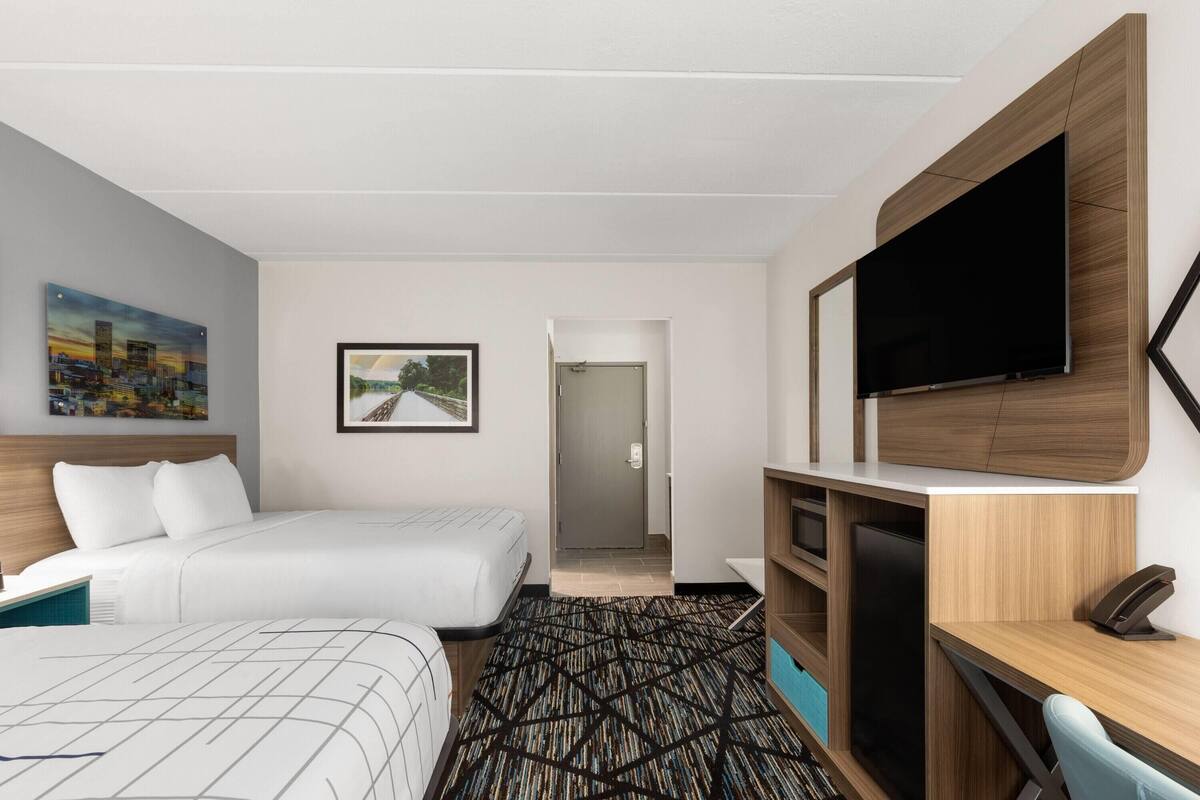 A hotel room with two beds, a desk with a flat-screen TV, a bookshelf, and a door leading to a hallway.