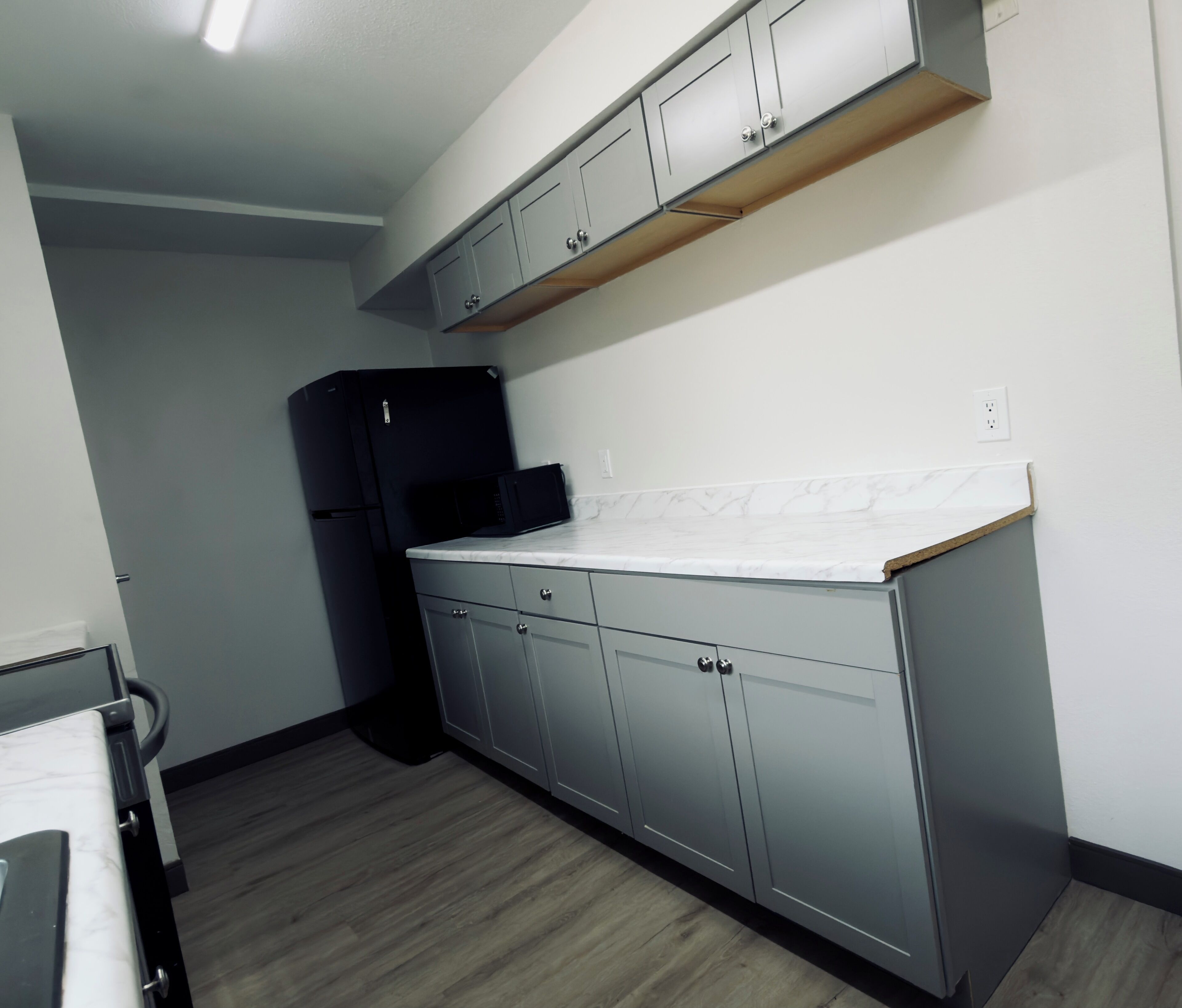 Eine Küche mit grauen Schränken, einem schwarzen Kühlschrank und einer weißen Arbeitsplatte.