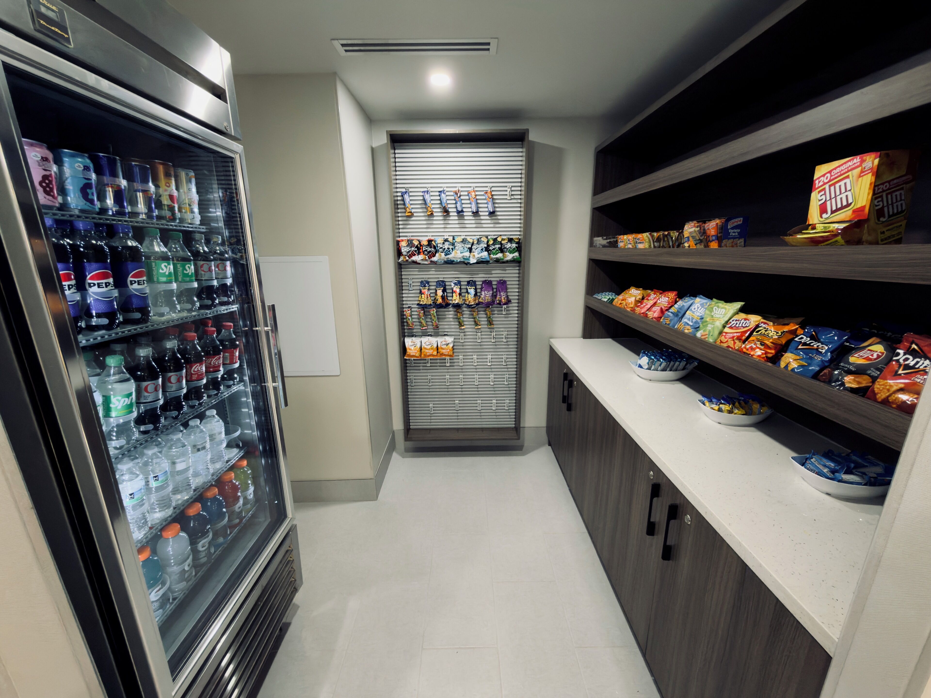 Un pasillo estrecho con un mostrador refrigerado de bebidas a la izquierda, una máquina expendedora en el centro y estantes con snacks a la derecha.