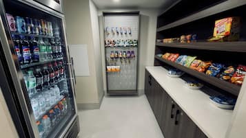 Un pasillo estrecho con un mostrador refrigerado de bebidas a la izquierda, una máquina expendedora en el centro y estantes con snacks a la derecha.
