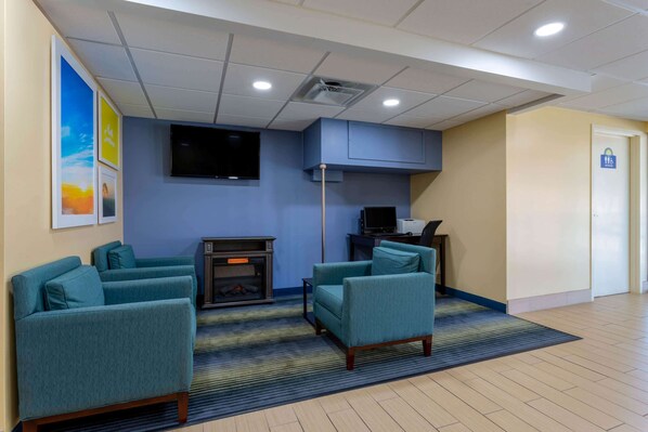 A hotel lobby with teal armchairs, a flat-screen TV, a fireplace, and a door marked with a wheelchair symbol.