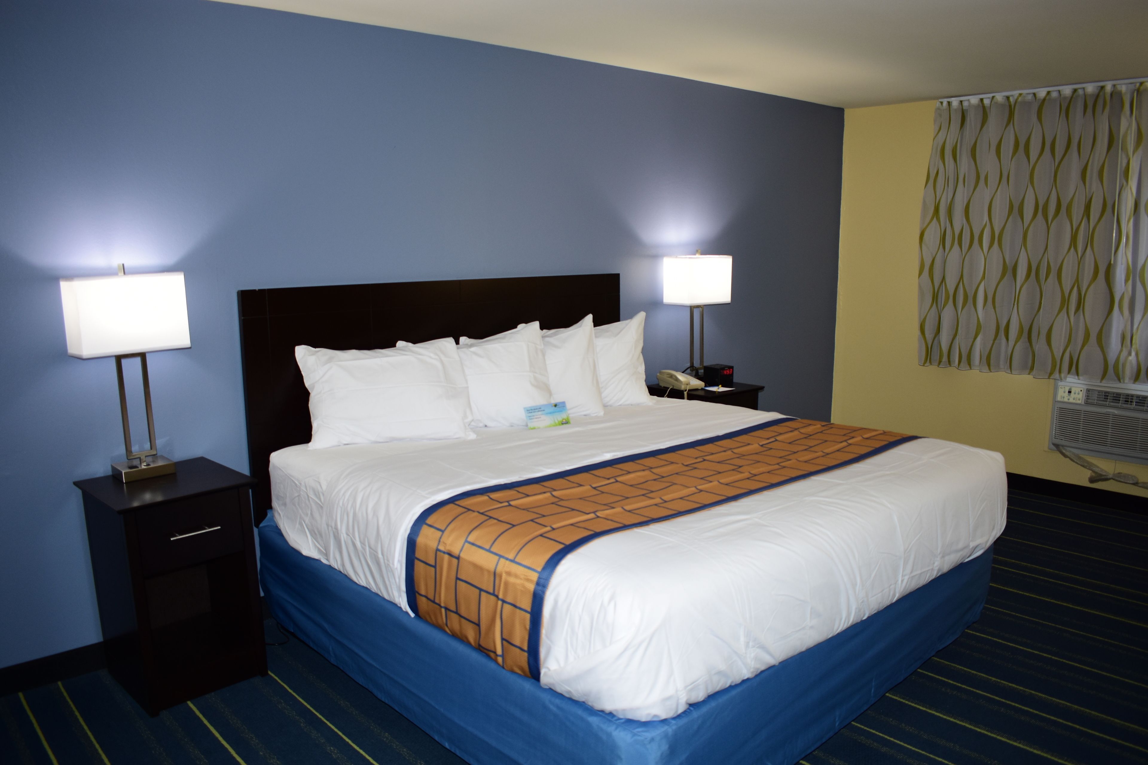 A hotel room with a large bed, two bedside tables with lamps, a wall-mounted air conditioner, and a patterned curtain.