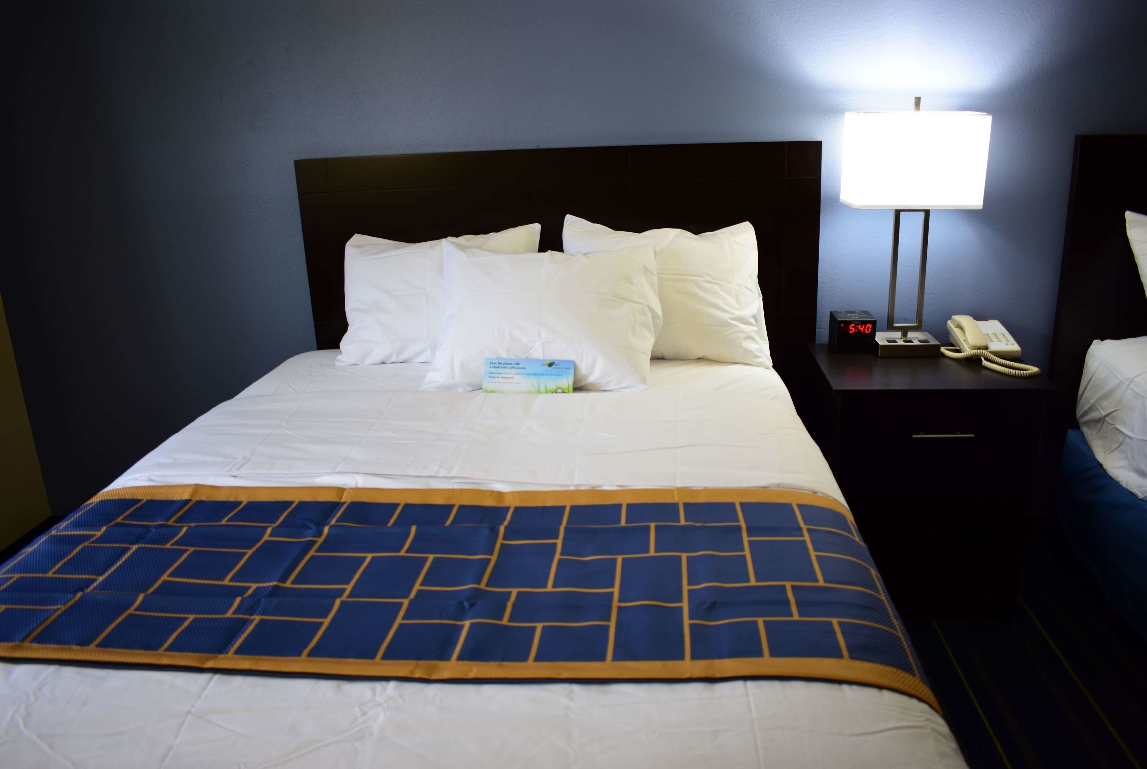 A neatly made bed with white linens and a blue and yellow patterned throw blanket. There is a bedside table with a lamp, a clock, and a telephone.