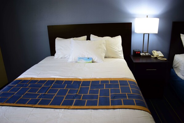 A neatly made bed with white linens and a blue and yellow patterned throw blanket. There is a bedside table with a lamp, a clock, and a telephone.