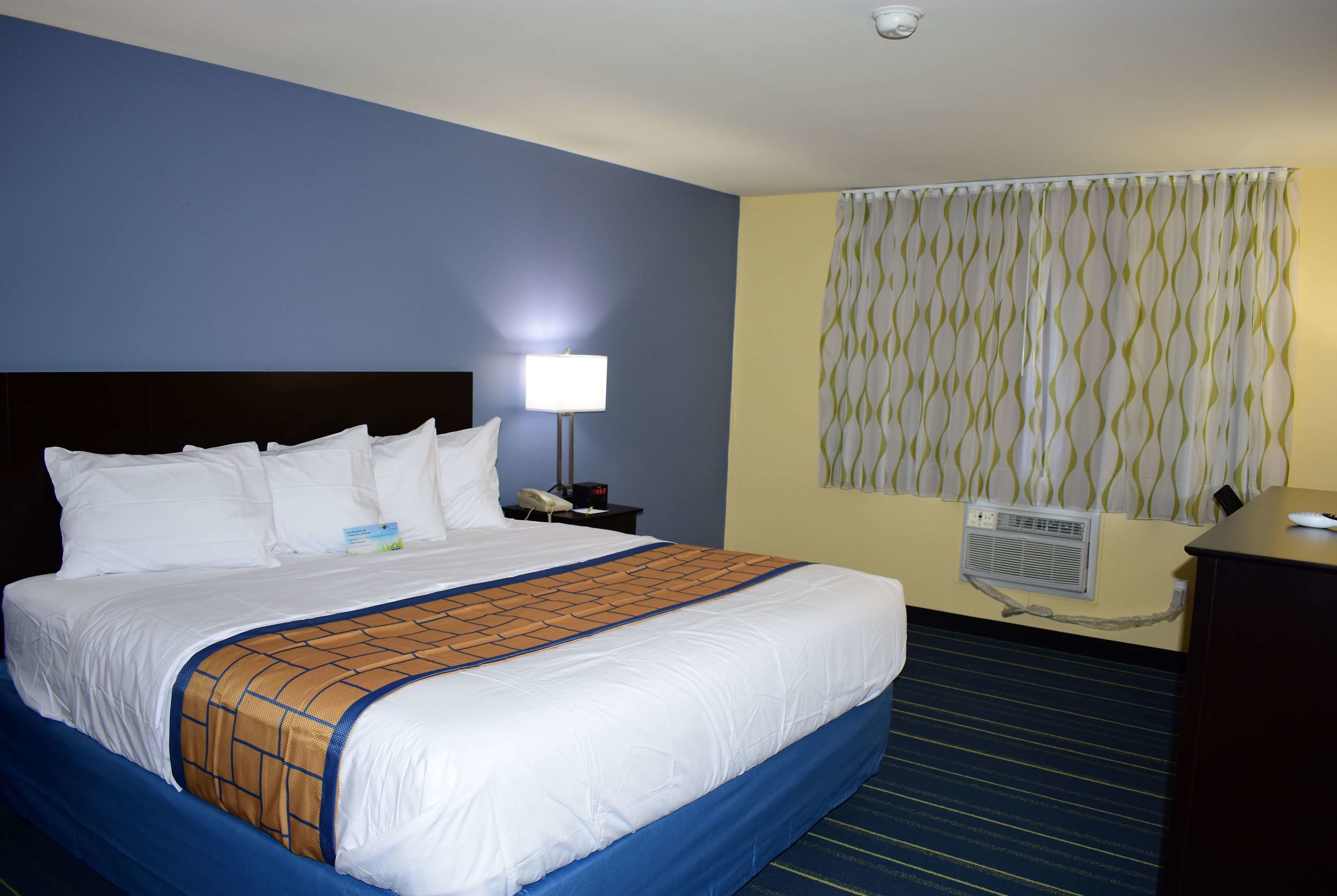 Habitación de hotel con una cama grande, una lámpara de noche, un teléfono, un equipo de aire acondicionado y una ventana con cortinas.