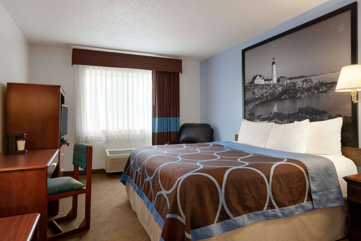 A hotel room with a bed, a desk, a chair, a television, and a painting of a lighthouse.