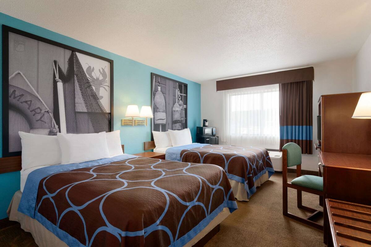 A hotel room with two beds, a desk, a chair, a TV, and artwork on the walls.
