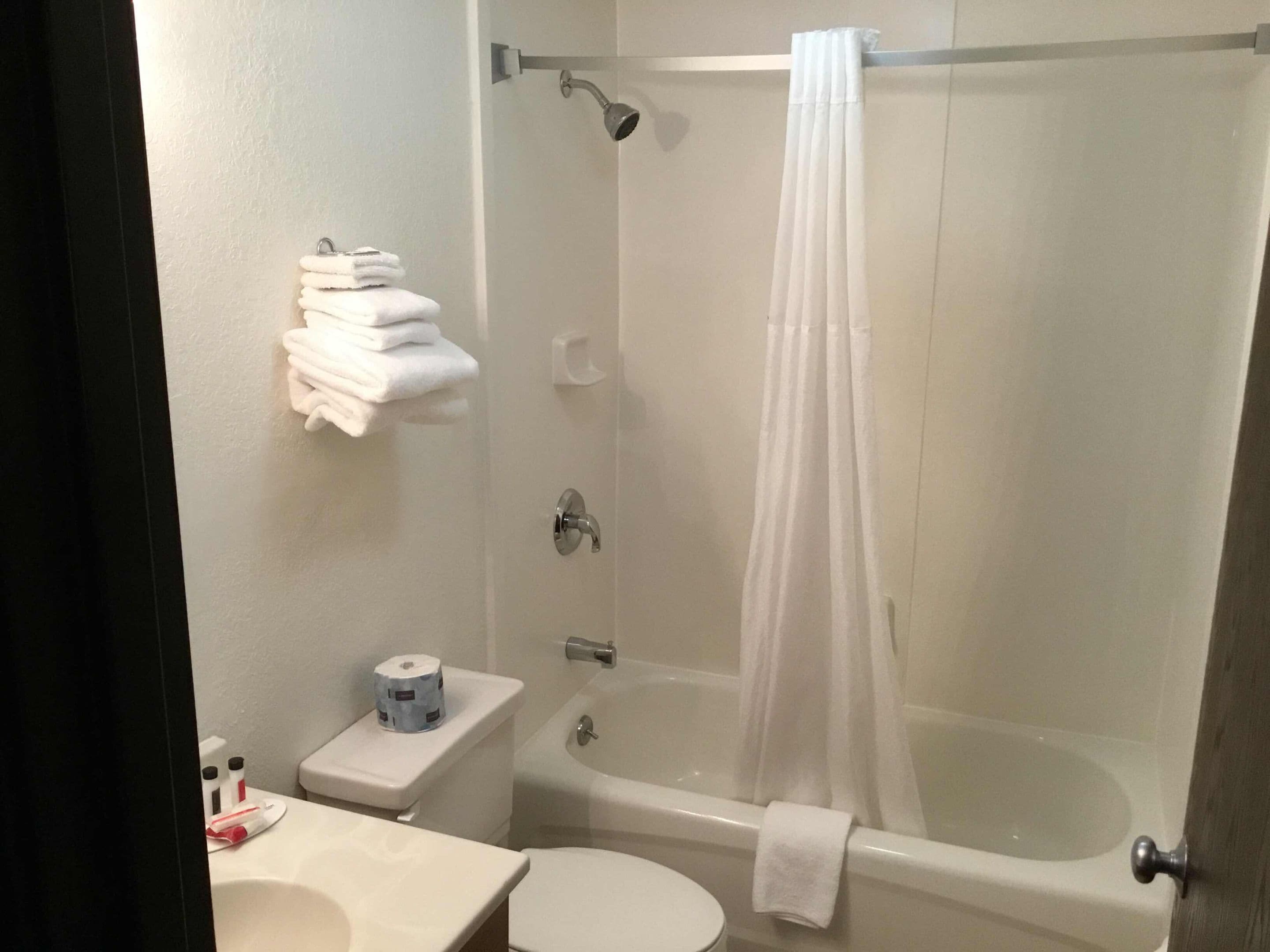 A bathroom with a bathtub, toilet, and sink. There are towels stacked on a shelf and a roll of toilet paper on the counter.