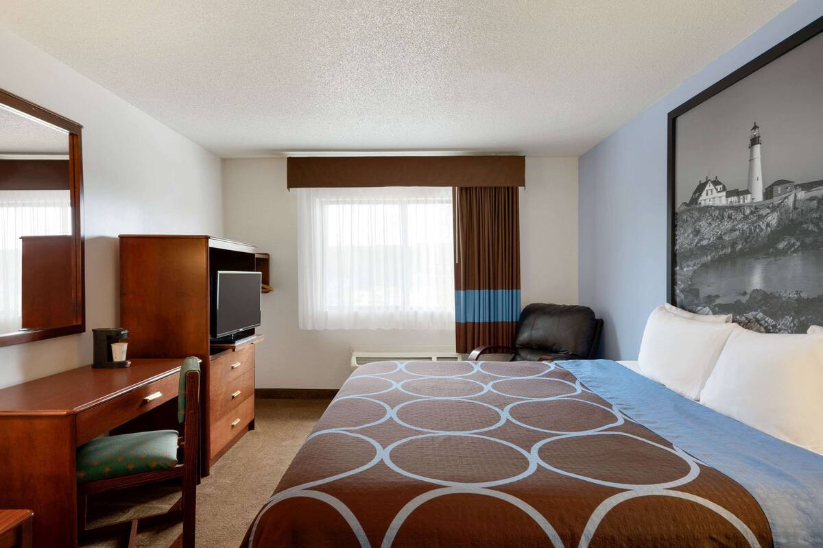 A hotel room with a bed, a desk, a chair, a television, and a painting of a lighthouse.