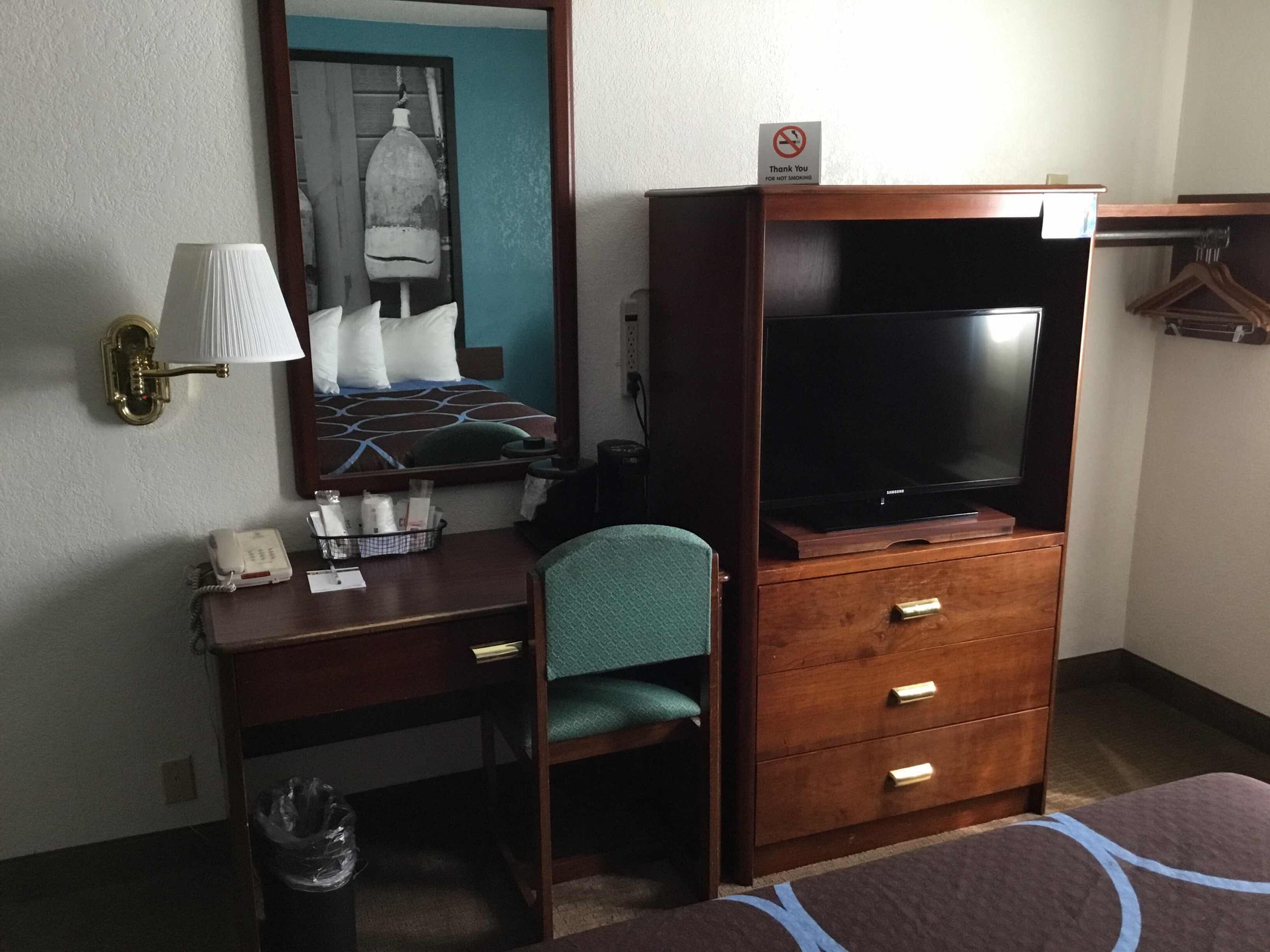 A hotel room with a wooden desk, a television, a chair, a trash bin, and a mirror.