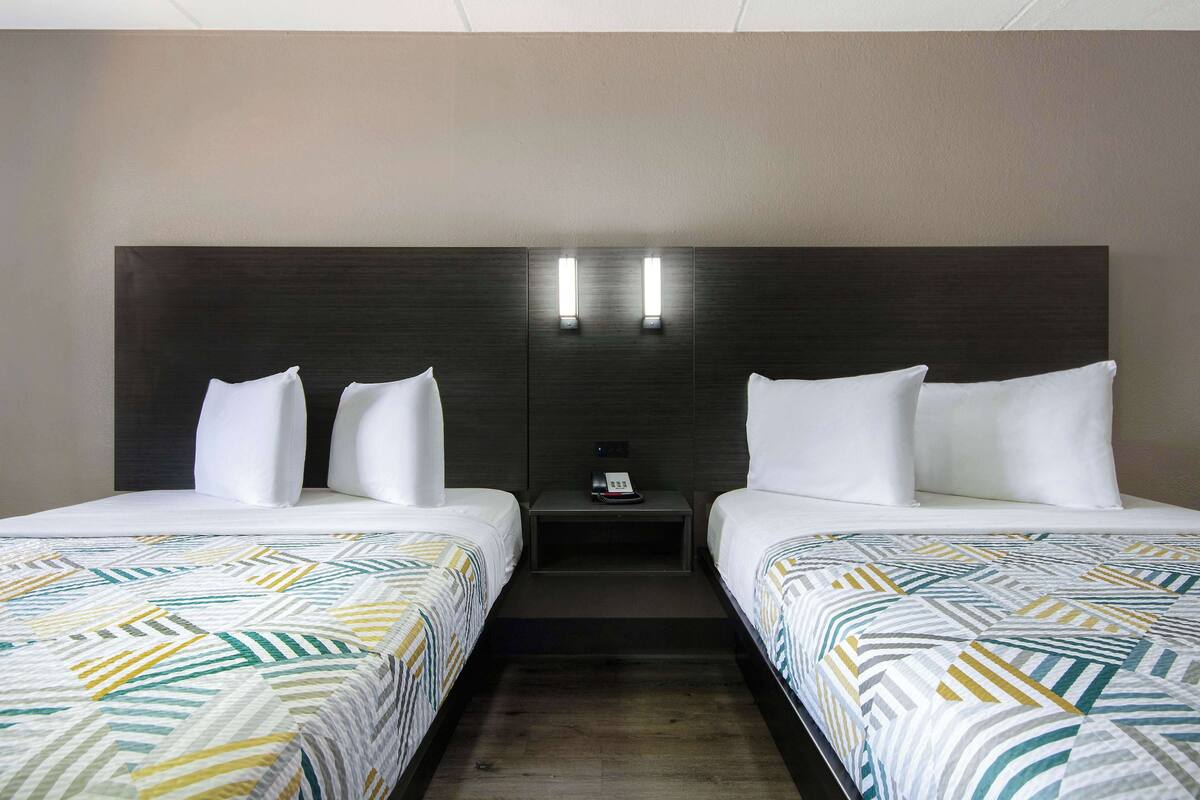 Une chambre d’hôtel avec deux lits, chacun doté de draps blancs et d’une couette à motifs géométriques. Il y a deux tables de chevet avec des lampes, et un téléphone sur l’une d’elles. La chambre est équipée d’une tête de lit en bois foncé et d’un mur aux couleurs neutres.