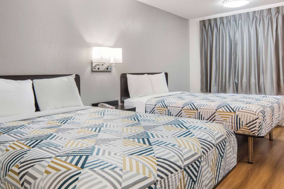 A hotel room with two beds, a nightstand with a phone, a wall-mounted lamp, and a window with curtains.