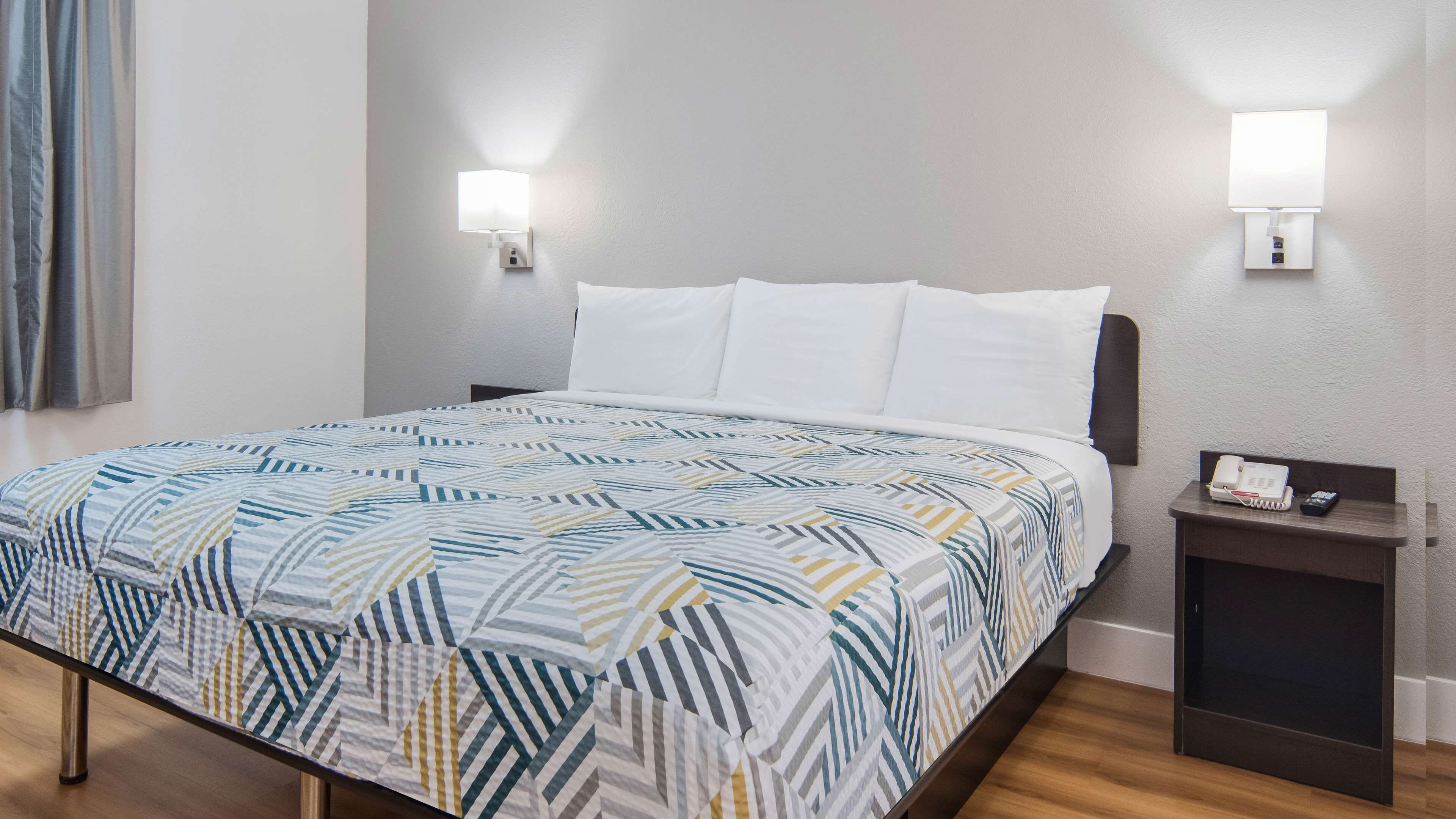 A neatly made bed with a geometric patterned bedspread, a bedside table with a phone and remote, and two wall-mounted lamps.