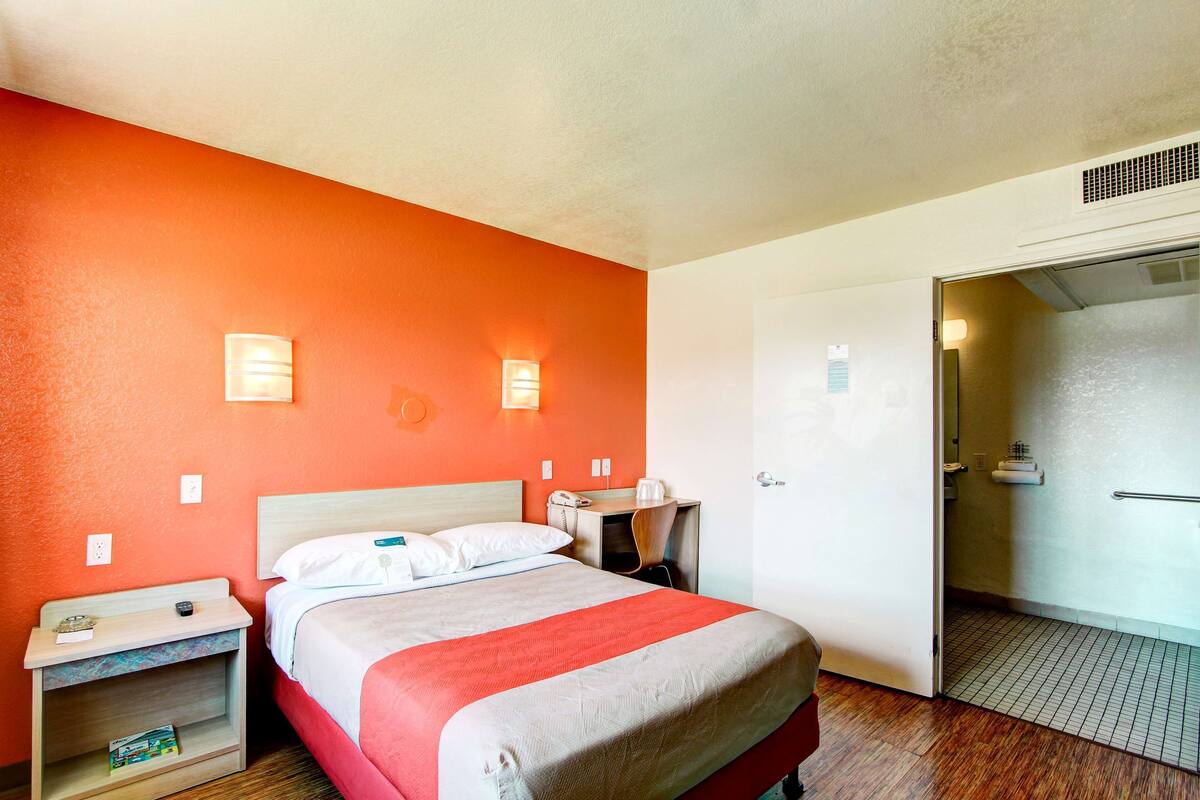 A hotel room with a bed, a nightstand, a desk, and a bathroom visible through an open door.