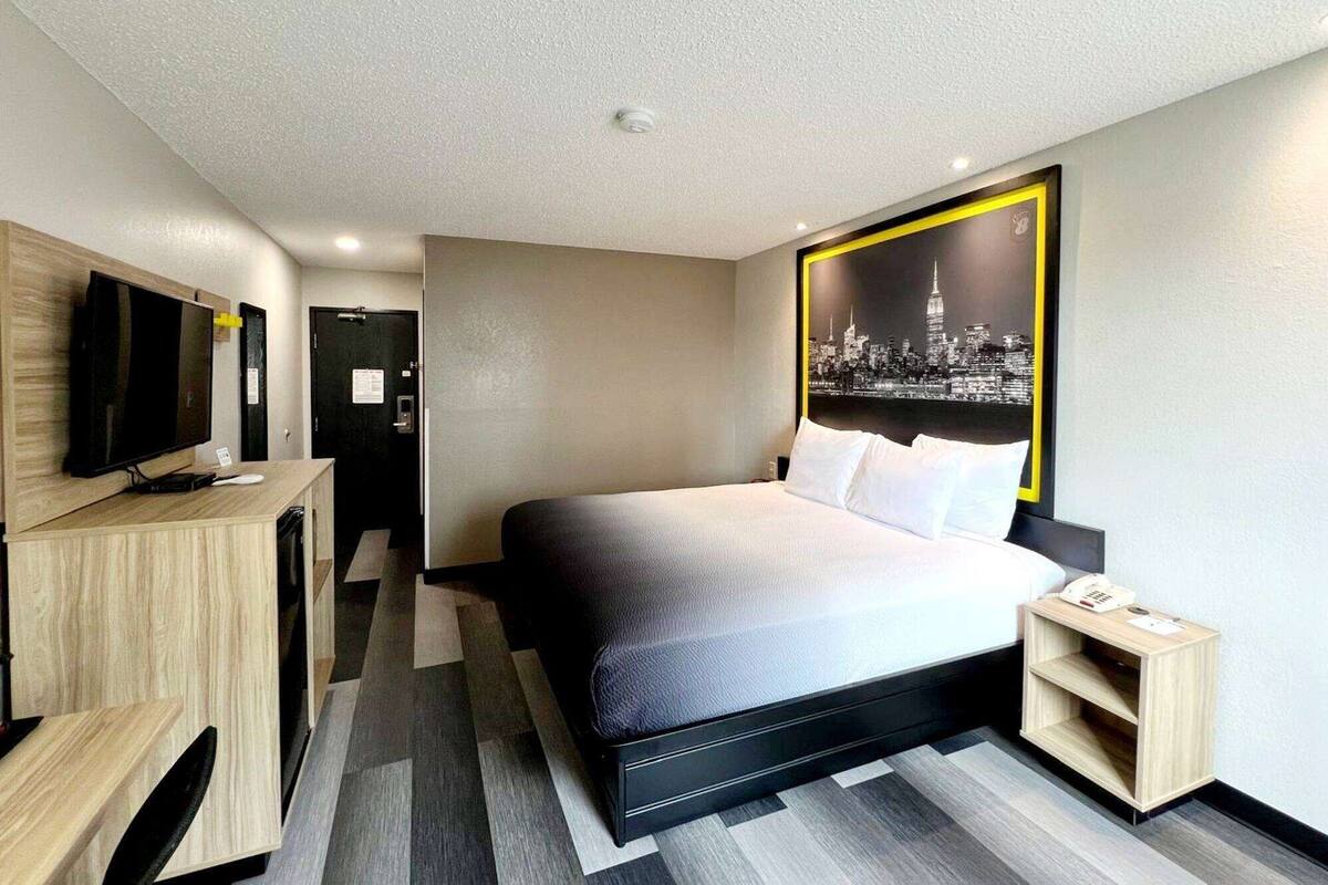Una habitación de hotel moderna con una cama grande, un televisor sobre un mueble de madera, un cuadro de un paisaje urbano en la pared y una mesita de noche pequeña.