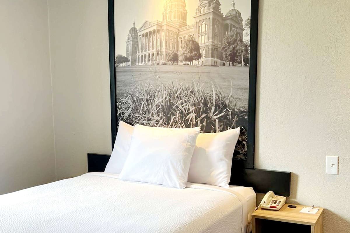 Habitación de hotel con cama, almohadillas blancas, mesita de noche con teléfono y un cuadro con la imagen de un edificio en la pared.