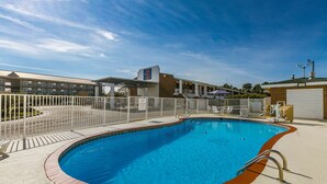 Una piscina con valla blanca, zona de estar techada y un edificio con un letrero que dice '6' al fondo.