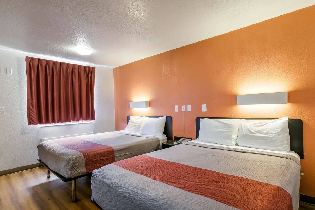 A hotel room with two beds, each with a headboard, a window with red curtains, and wall-mounted lights.