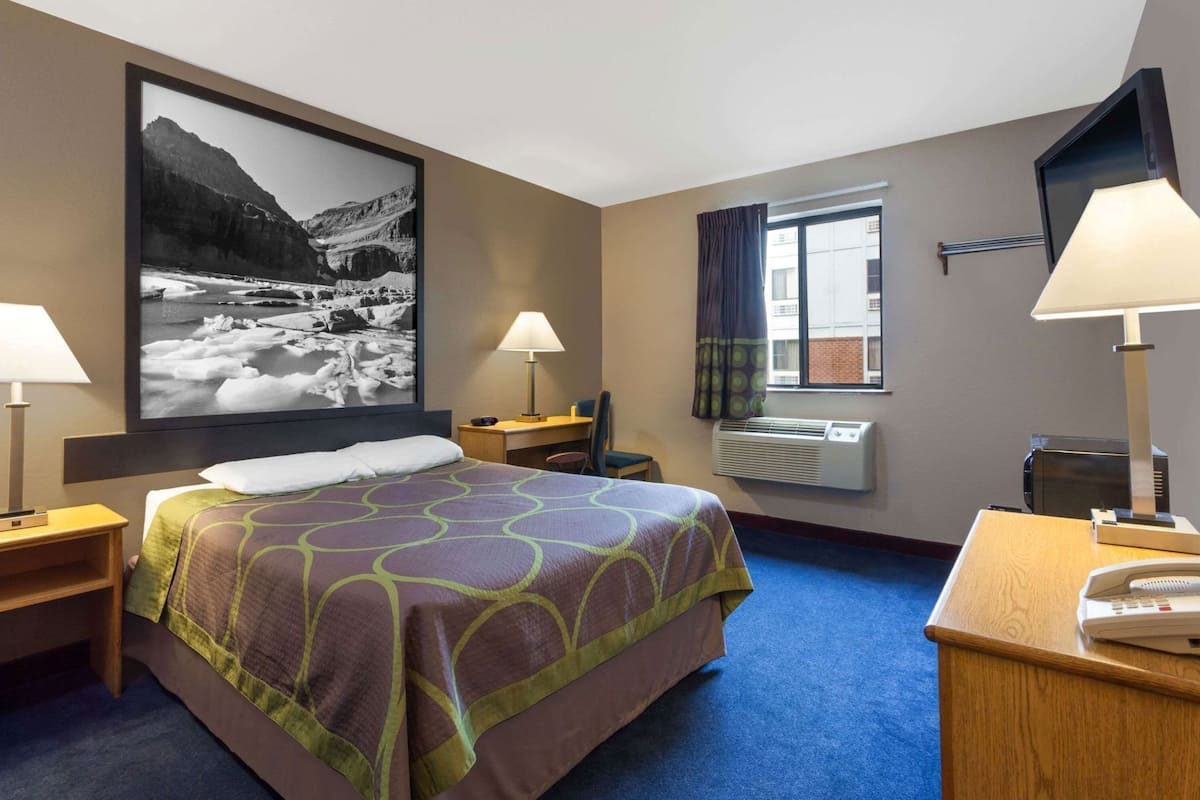 A hotel room with a bed, two bedside tables, a desk, a chair, a large window with curtains, an air conditioning unit, and a television.