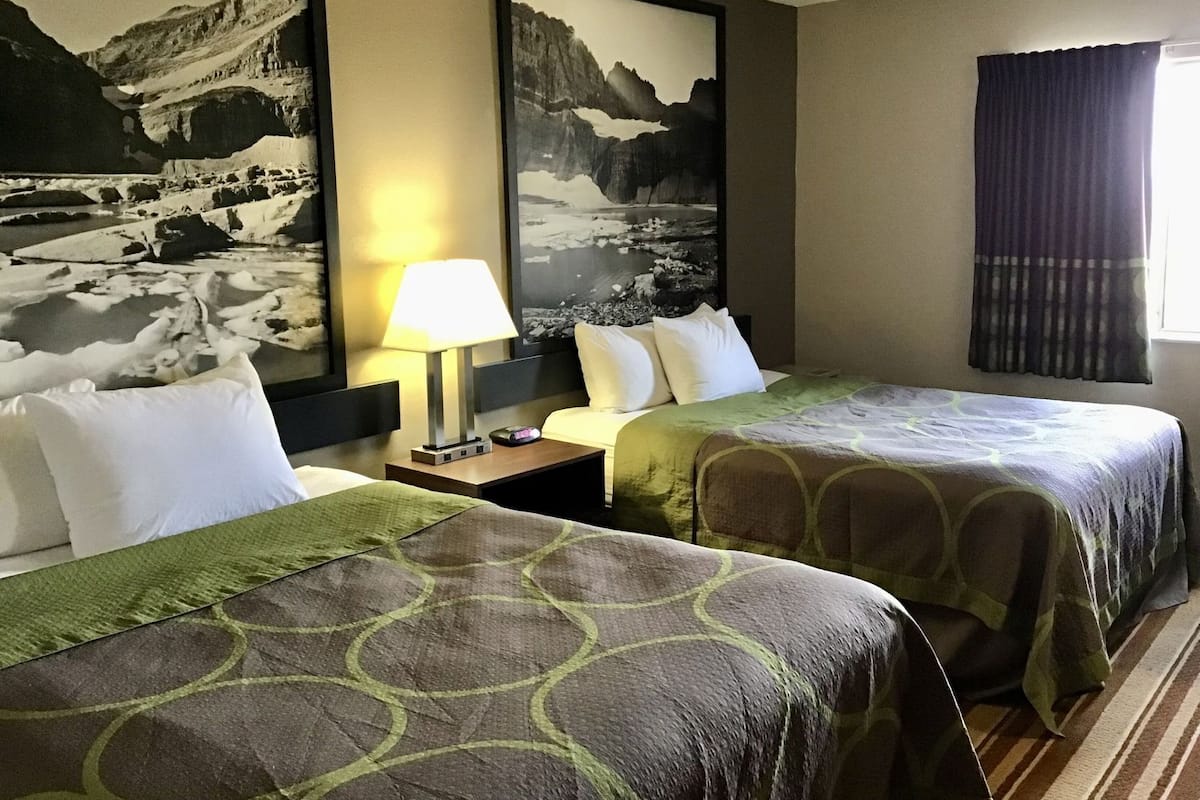 A hotel room with two beds, a nightstand with a lamp, and two large framed pictures of mountains.